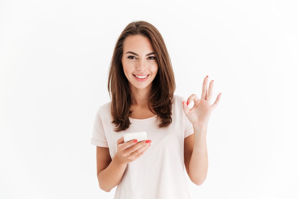 Carefree brunette woman holding smartphone and showing ok sign